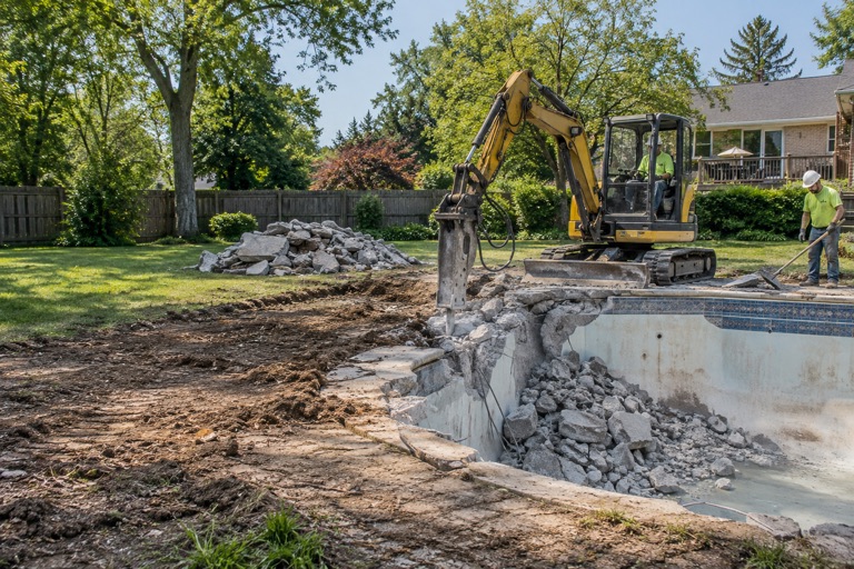Excavator with a hydraulic breaker cracking apart an inground pool shell and concrete deck in a Peoria, Illinois backyard